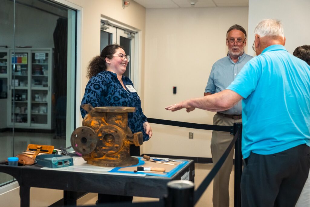 Two people on the outside of a rope speak to a person on the other side of the rope, standing next to a table that holds a rusted artifact