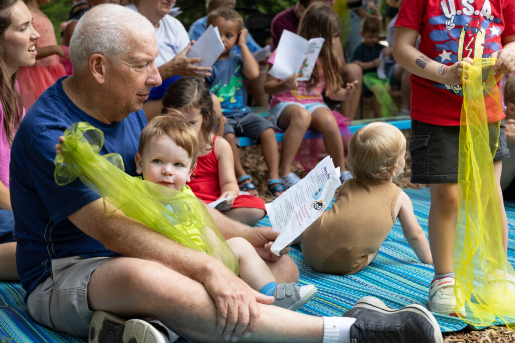 Grandfather with grandson at Wonder Wednesday. Grandfather is following along with the songbook while the grandson plays with a scarf. There are other children and caregivers in the background.