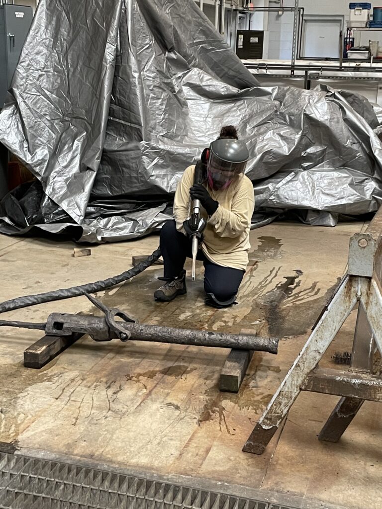 A person kneels next to a small cannon, holding the nozzle to a dry ice blasting apparatus shaped like a long, thin tapering rectangle with a hose that extends off-screen. A tarp covers the background, with a sawhorse to the side of the person.