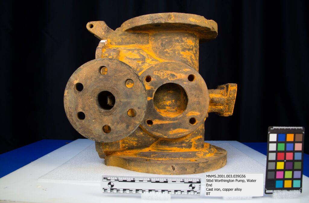A rust-covered component of a pump sits at the center of the image with a black background, on A blue table covered in white foam. A scale bar, label, and color card sit in the foreground.