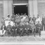 A black and white photo of a group of people sitting and standing in front of a building entrance wearing formal and nursing attire