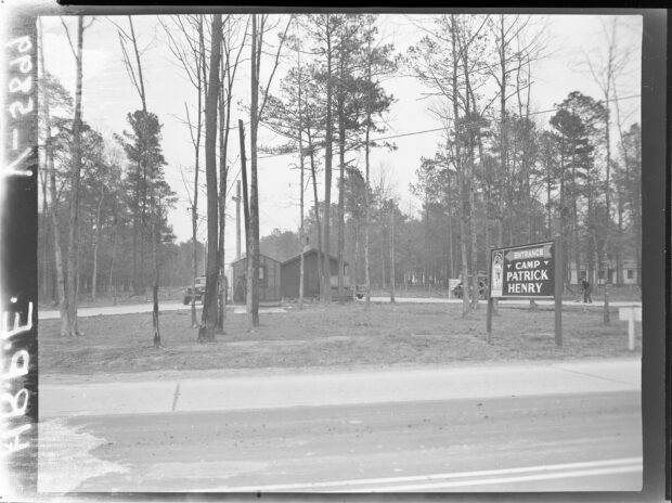black and white photo of a building behind trees with the sign "Camp Patrick Henry" by the road