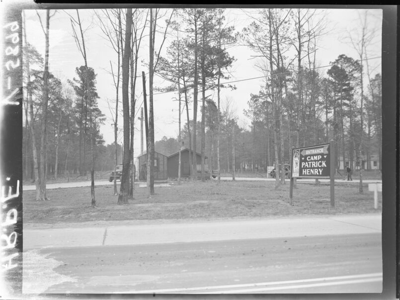 black and white photo of a building behind trees with the sign "Camp Patrick Henry" by the road