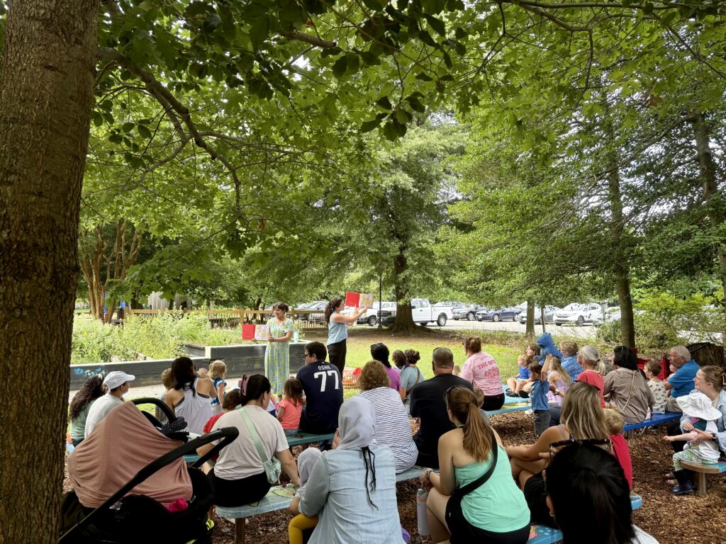 Caregivers, children, and educators at Wonder Wednesday. Educators are reading a book to the crowd. The group is outside in a picnic/garden area.