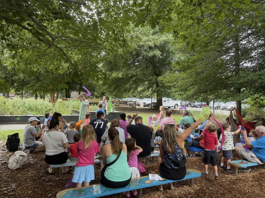 Caregivers, children, and educators singing and dancing during Wonder Wednesday. The crowd is outside in a picnic/garden area.