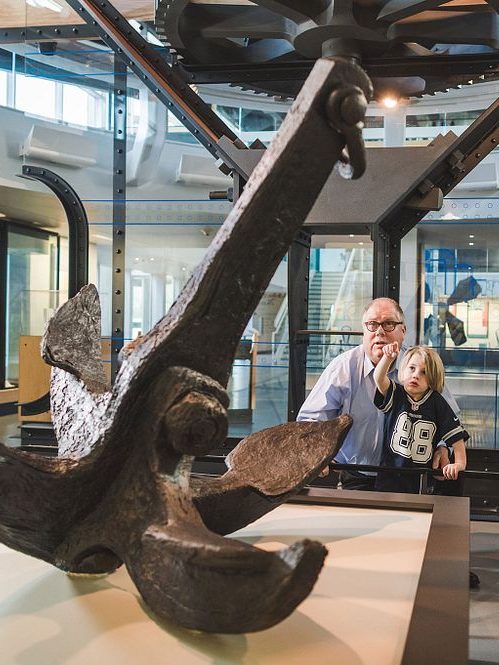 Grandfather and grandson looking at USS Monitor's conserved anchor in the large artifact gallery in the USS Monitor Center. The little boy is pointing at the anchor while the grandfather has his arm around him.